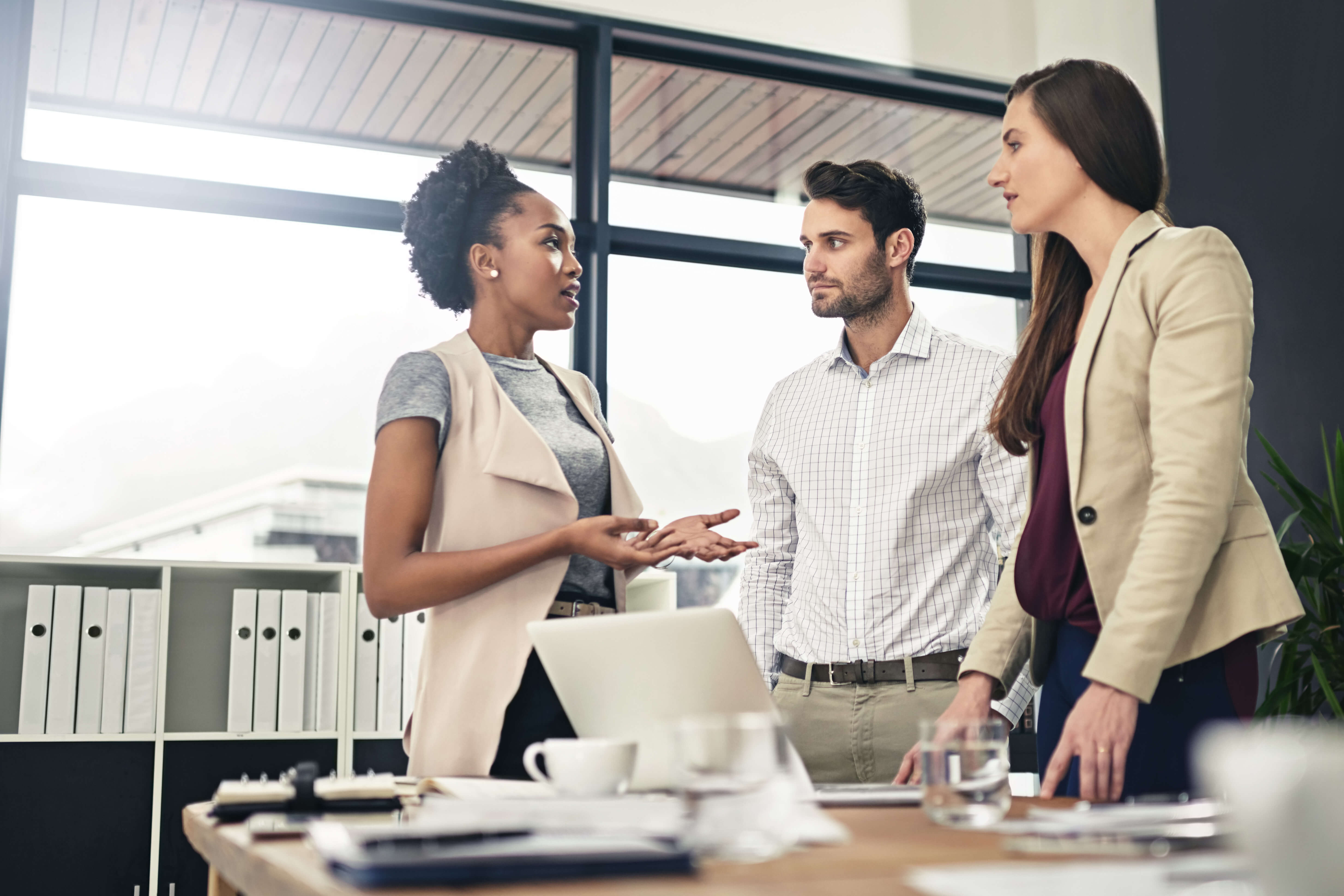 Three professionals having a discussion in a modern office, standing near a desk with documents, a laptop, and coffee cups, emphasizing teamwork and collaboration